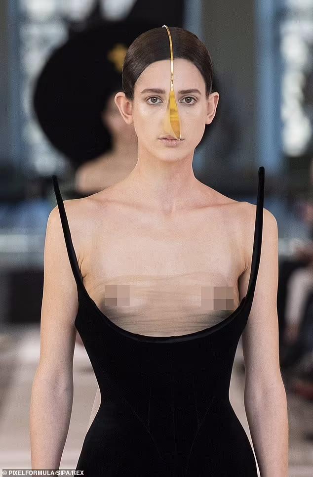 Một người mẫu mặc váy đen khoét cổ rộng lộ rõ vòng một sau lớp vải xuyên thấu "có như không". Ngoài ra, món đồ trang sức gắn trên đầu kéo dài xuống mũi trông khó hiểu.