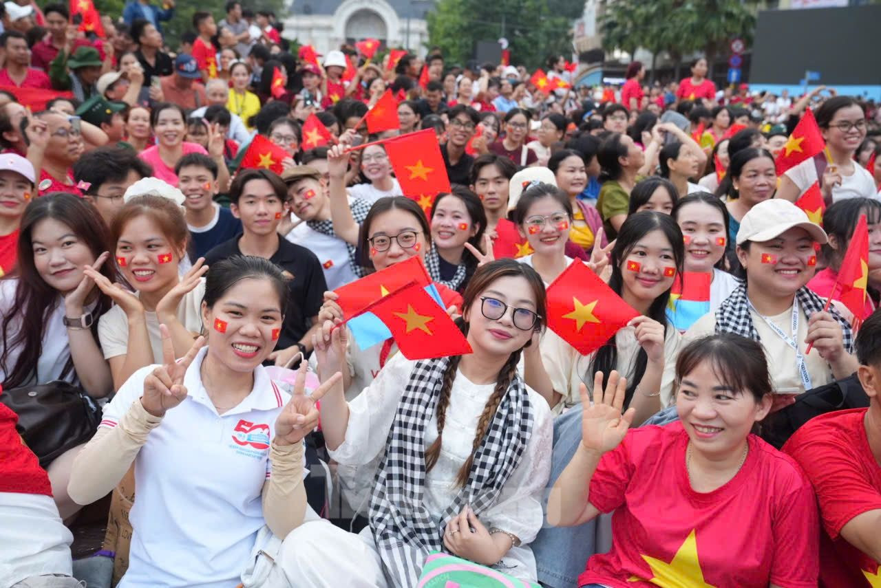 Bên cạnh cụ là những thanh thiếu niên rạng rỡ trong áo cờ đỏ, không ngừng reo vang lời ca quen thuộc: Như có Bác Hồ trong ngày vui đại thắng, Tiến về Sài Gòn, Bác đang cùng chúng cháu hành quân... Những giai điệu ấy hòa trong tiếng trống, tiếng nhạc quân hành, tạo nên không gian lễ hội vừa hùng tráng vừa đậm chất dân tộc.