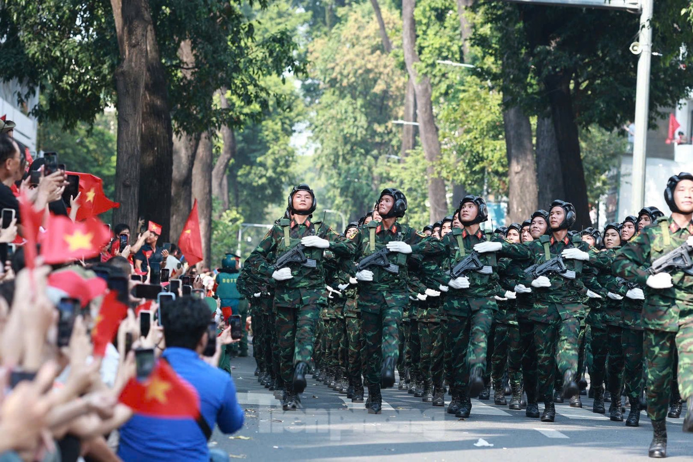 Bên cạnh cụ là những thanh thiếu niên rạng rỡ trong áo cờ đỏ, không ngừng reo vang lời ca quen thuộc: Như có Bác Hồ trong ngày vui đại thắng, Tiến về Sài Gòn, Bác đang cùng chúng cháu hành quân... Những giai điệu ấy hòa trong tiếng trống, tiếng nhạc quân hành, tạo nên không gian lễ hội vừa hùng tráng vừa đậm chất dân tộc.