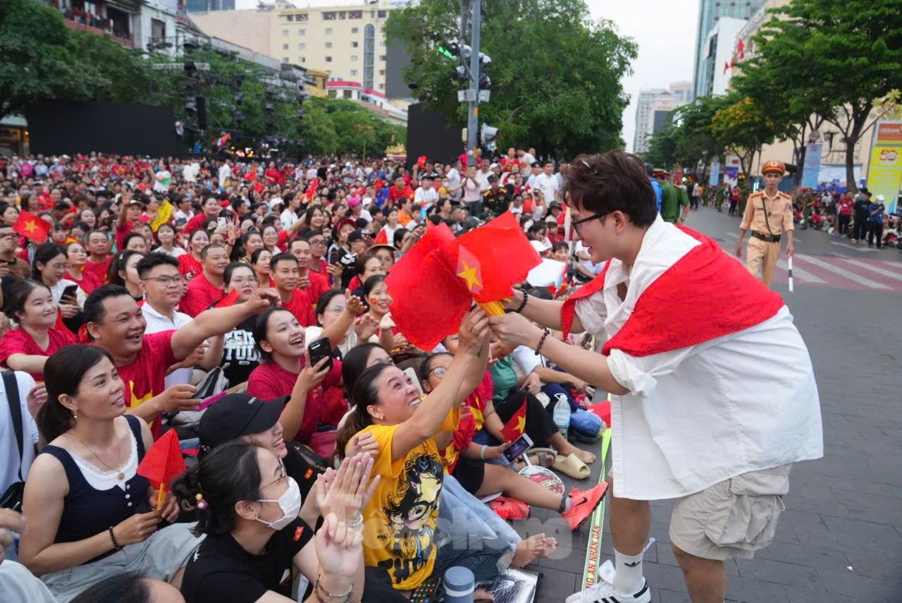 Bên cạnh cụ là những thanh thiếu niên rạng rỡ trong áo cờ đỏ, không ngừng reo vang lời ca quen thuộc: Như có Bác Hồ trong ngày vui đại thắng, Tiến về Sài Gòn, Bác đang cùng chúng cháu hành quân... Những giai điệu ấy hòa trong tiếng trống, tiếng nhạc quân hành, tạo nên không gian lễ hội vừa hùng tráng vừa đậm chất dân tộc.