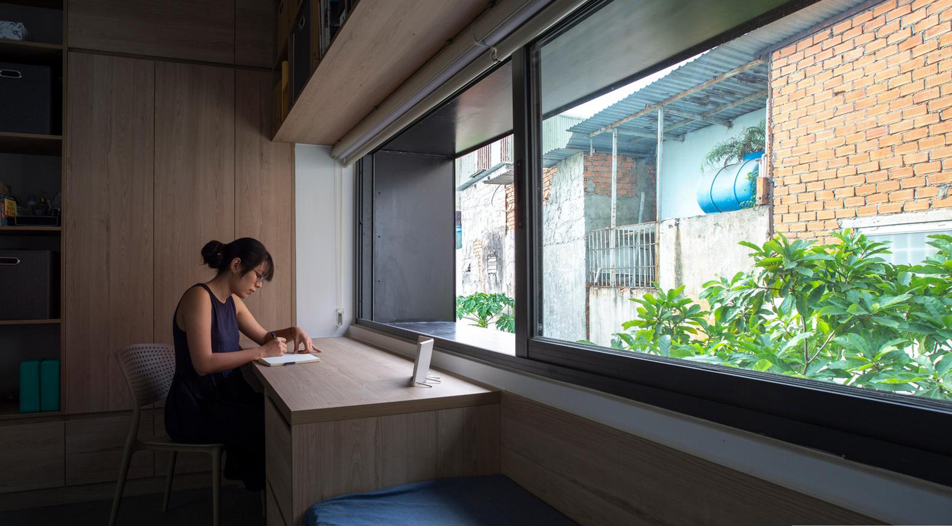 Thành phố Hồ Chí Minh là một trong những đô thị đông dân nhất của Đông Nam Á nên cuộc sống vô cùng bộn bề, ngột ngạt cũng như tù túng. Hơn nữa, thành phố này cũng đang diễn ra làn sóng COVID -19 lần thứ 5 vô cùng nguy hiểm với hàng nghìn ca mắc mỗi ngày. Do đó mọi người thường phải làm việc online tại nhà, nếu không muốn nói là bị “mắc kẹt” ở nhà. Vì thế một nhóm kiến trúc sư đã thực hiện ý tưởng làm ngôi nhà với không gian ở thành nơi làm việc để ứng phó với đại dịch. Các kiến trúc sư mong muốn ngôi nhà giúp các thành viên trong gia đình có cơ hội sống chậm lại để trải nghiệm không gian. Đồng thời nó phải đảm bảo tiết kiệm ngân sách với giá cả phải chăng và thi công trong thời gian ngắn nên thiết kế cực kỳ tối giản và đơn giản. Trong nhà, không có ranh giới cụ thể giữa các không gian vì phòng bếp chính là phòng ăn, phòng ngủ cũng là nơi làm việc. Việc sử dụng kết hợp không gian được thể hiện dọc theo hành lang chính, nơi có một kệ tủ lớn và dài 14 mét gắn vào tường. Chiếc kệ này kéo dài, được sử dụng cho nhiều mục đích, là nơi để các vật dụng gia đình như nhà bếp, tủ đựng thức ăn, thiết bị, tủ lạnh, máy giặt, kệ giày dép, kệ sách,…Mọi thứ được sắp xếp và lấp đầy trong một cấu trúc có trật tự tạo nên sự đa dạng và linh hoạt. Không giống như nhà phố điển hình ở TP Hồ Chí Minh, ngôi nhà không mở ra bên ngoài mà hướng vào bên trong, Hai mặt tiền đơn giản hết sức có thể vì cả mặt trước và mặt sau chỉ có một khoảng hở “vừa đủ” đón ánh sáng ban ngày và thông gió, tránh ồn và ô nhiễm không khí cũng như duy trì sự riêng tư và thân mật của gia chủ. Ngôi nhà dành cho người hướng nội nên được thiết kế để nhìn từ trong ra ngoài và nhận được năng lượng từ sự phản chiếu bên trong. Các cửa trượt đều làm bằng kính để tăng sự rộng rãi cho không gian cũng như ánh sáng cho cả ngôi nhà. Cầu thang và nhà vệ sinh được bố trí ở phía trước nhà, sân trong nằm ở trung tâm của ngôi nhà. Còn bếp, phòng khách và các phòng ngủ được bố trí phía sau ở hai bên sân trong, tạo sự thông thoáng và đón nhận ánh sáng cho cả ngôi nhà.