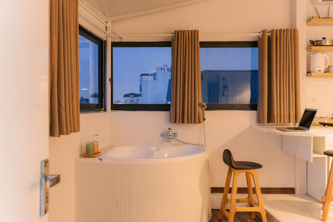 Với chi phí 150 triệu đồng, Tùng Anh hài lòng với căn "mini penthouse" của mình. Sau hơn nửa tháng thiết kế và thi công, sân thượng trở thành phòng riêng của cậu chủ trẻ. Phòng ở ngoài "view" hai mặt phố cổ còn có đầy đủ nội thất và tiện nghi sinh hoạt. Gia chủ bố trí hệ tivi thông minh với tay nâng và xoay đa chiều để ngồi ở sofa bệt hoặc nằm trên giường đều xem được mà vẫn nhìn ra phố cổ. Góc thư giãn ưa thích nhất của Tùng Anh là bồn tắm. Thời gian hoàn thiện bồn tắm chiếm hơn nửa tổng quá trình cải tạo. Bồn tắm ốp gạch thẻ que Mosaic. Gia chủ sử dụng keo Saveto của Italy thay cho xi măng trắng truyền thống để chít mạch gạch. Góc bếp và quầy bar nhỏ bởi gia chủ là người ít làm bếp nhưng vẫn đủ nấu các món đơn giản. Bàn quầy bar có thể gập xuống bằng mặt bếp. Ngăn tủ mini bên dưới tăng không gian để đồ và đỡ mặt bàn. Quầy bar kiêm bàn làm việc và bàn ăn kia cũng có thể gập lại nhằm tiết kiệm diện tích sử dụng trong phòng. Đợt gỗ ở bếp được gắn đèn LED hắt sáng. Chiếc sofa lười đỡ tốn diện tích và dễ di chuyển. Gia chủ làm thêm đôi đèn treo để đọc sách và nghỉ ngơi buổi tối.