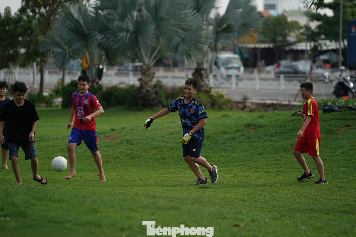 Một nhóm trẻ em nô đùa, chơi bóng trên thảm cỏ xanh mướt của một chung cư, phía sau là đoạn Nguyễn Văn Linh vừa được mở rộng.