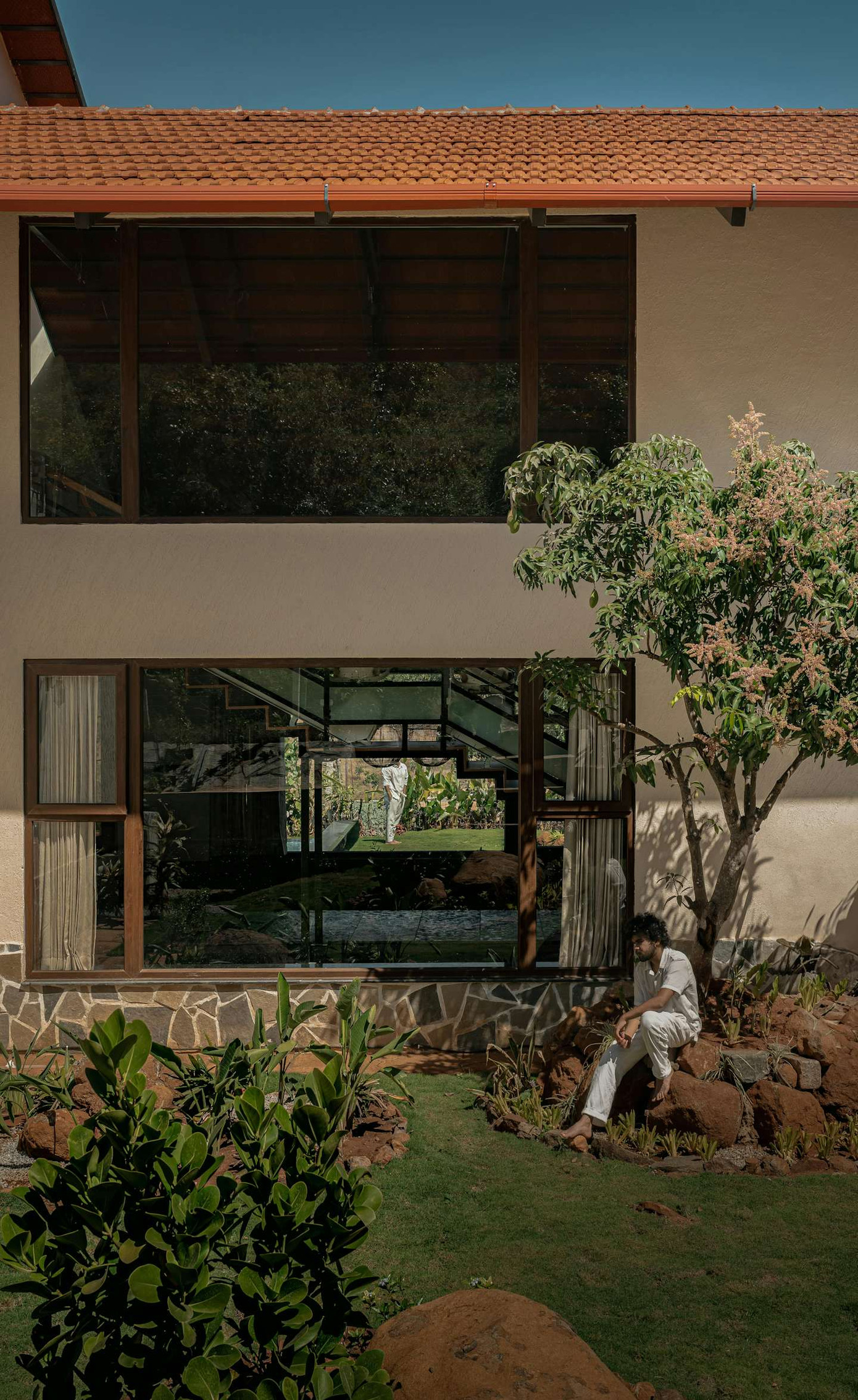 Một góc mặt đứng của ngôi nhà hai tầng với mái ngói đỏ và bức tường sơn màu be nhạt, mang lại phong cách kiến trúc nhiệt đới hiện đại.