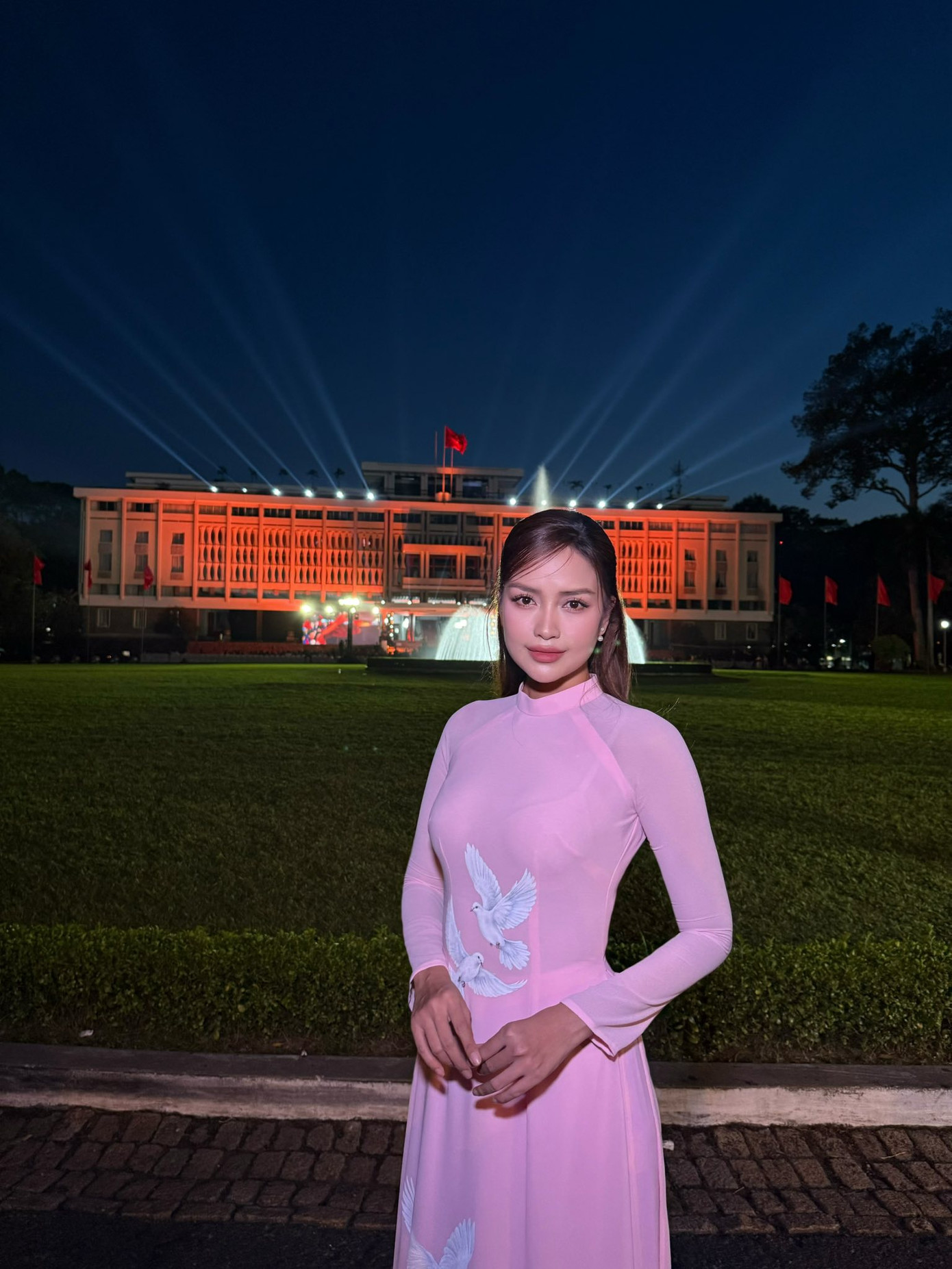 Hoa hậu Ngọc Châu mặc áo dài tông màu hồng, in họa tiết chim bồ câu trắng khi chụp ảnh trước Dinh Độc Lập.
