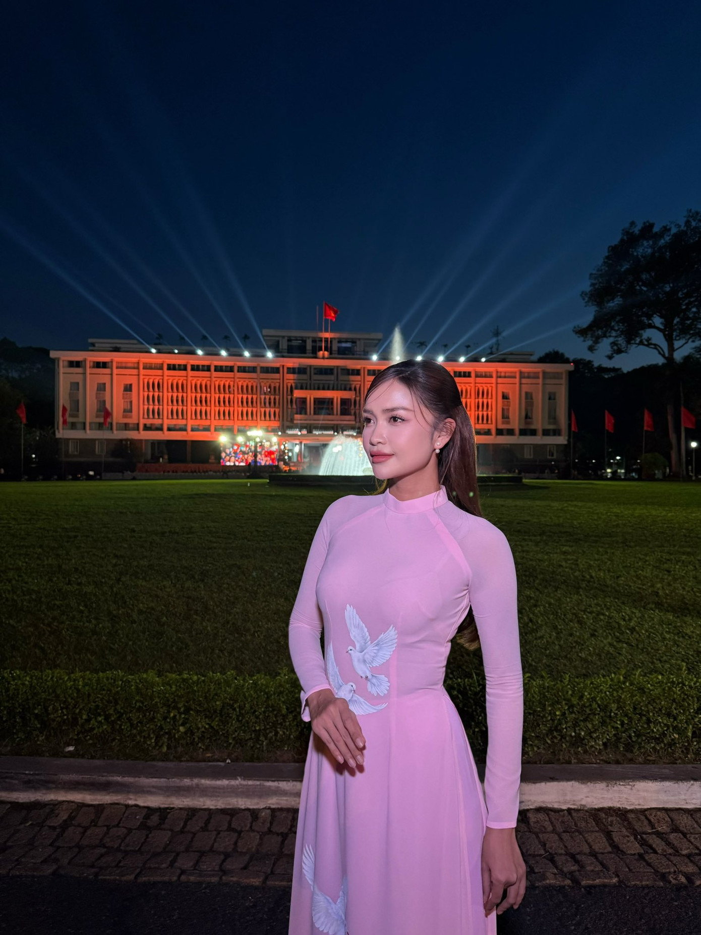 Hoa hậu Ngọc Châu mặc áo dài tông màu hồng, in họa tiết chim bồ câu trắng khi chụp ảnh trước Dinh Độc Lập.