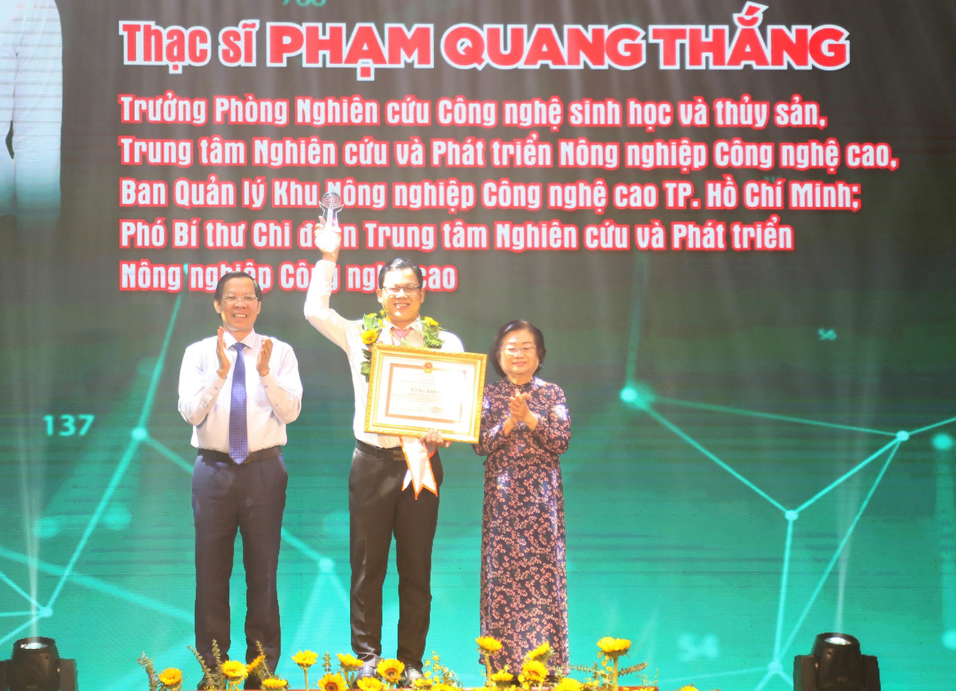 Bà Trương Mỹ Hoa, nguyên Phó Chủ tịch nước (bên phải); ông Phan Văn Mãi, Phó Bí thư Thường trực Thành ủy, Chủ tịch UBND TPHCM (bên trái) trao hoa và bằng khen tặng Thạc sĩ Phạm Quang Thắng, Trưởng Phòng Nghiên cứu Công nghệ sinh học và thủy sản, Trung tâm Nghiên cứu và Phát triển Nông nghiệp Công nghệ cao (Ban Quản lý Khu Nông nghiệp Công nghệ cao TPHCM).