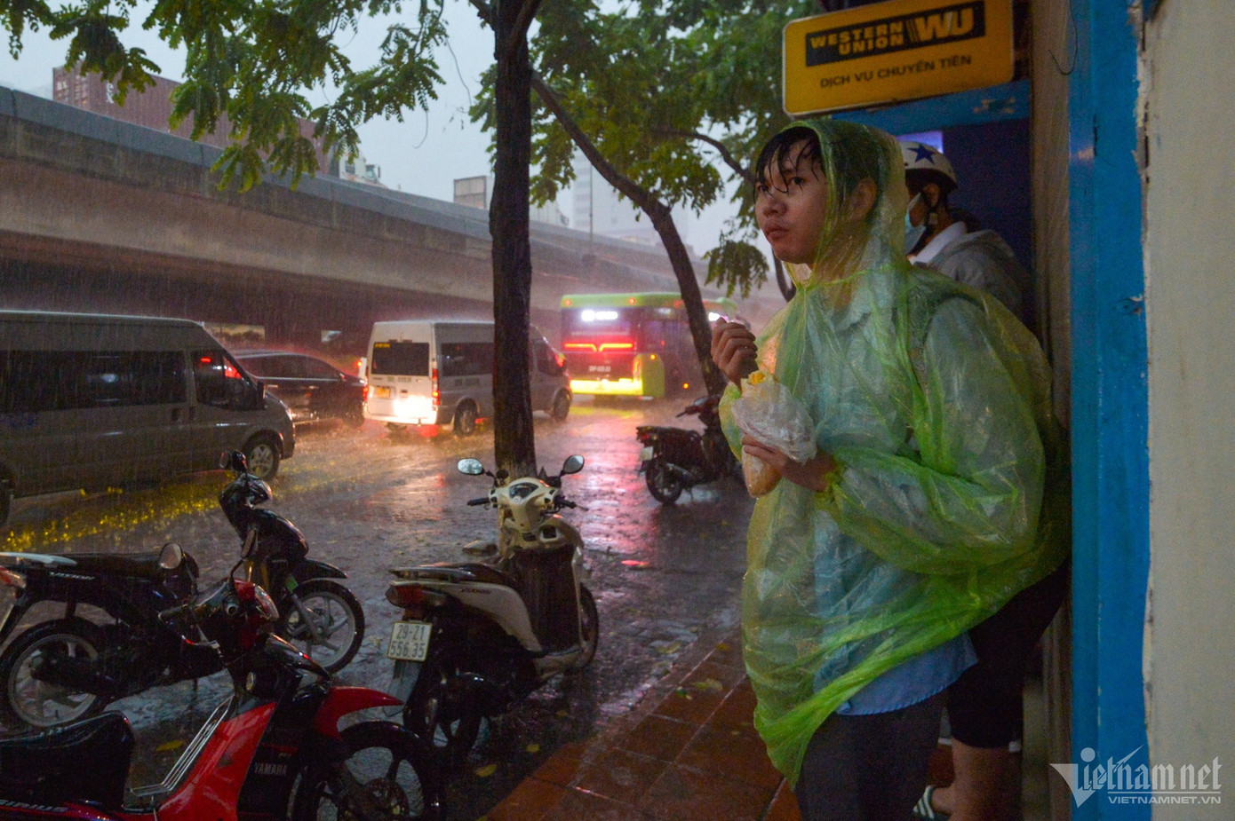 Thức dậy từ sớm, anh Long đi xe ôm từ nhà ra đường Khuất Duy Tiến mất 15 phút mới bắt được xe khách về quê. Trong khi chờ, nam thanh niên ăn vội ổ bánh mì dưới cơn mưa tầm tã.