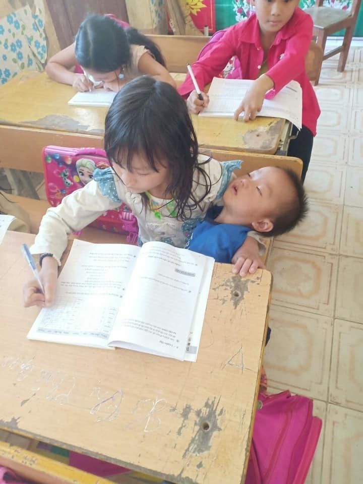 Cô bé vừa viết bài vừa bế em trên tay ngủ say.