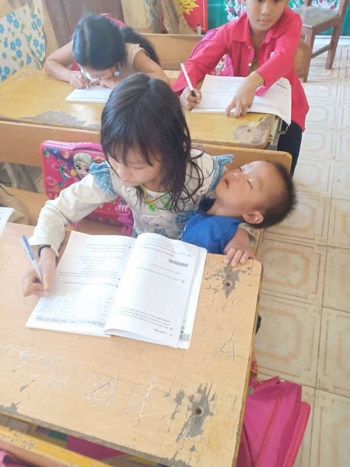 Cô bé vừa viết bài vừa bế em trên tay ngủ say.