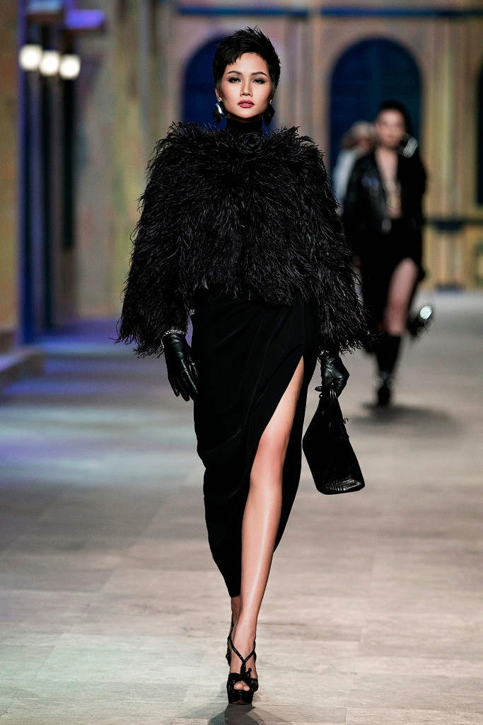 Người đẹp 26 tuổi sải những bước chân đầu tiên khuấy động không khí sàn diễn với bộ trang phục lấy sắc đen làm chủ đạo, kết hợp giữa chân váy xẻ tà bất đối xứng cùng áo cổ lọ, áo khoác lông.