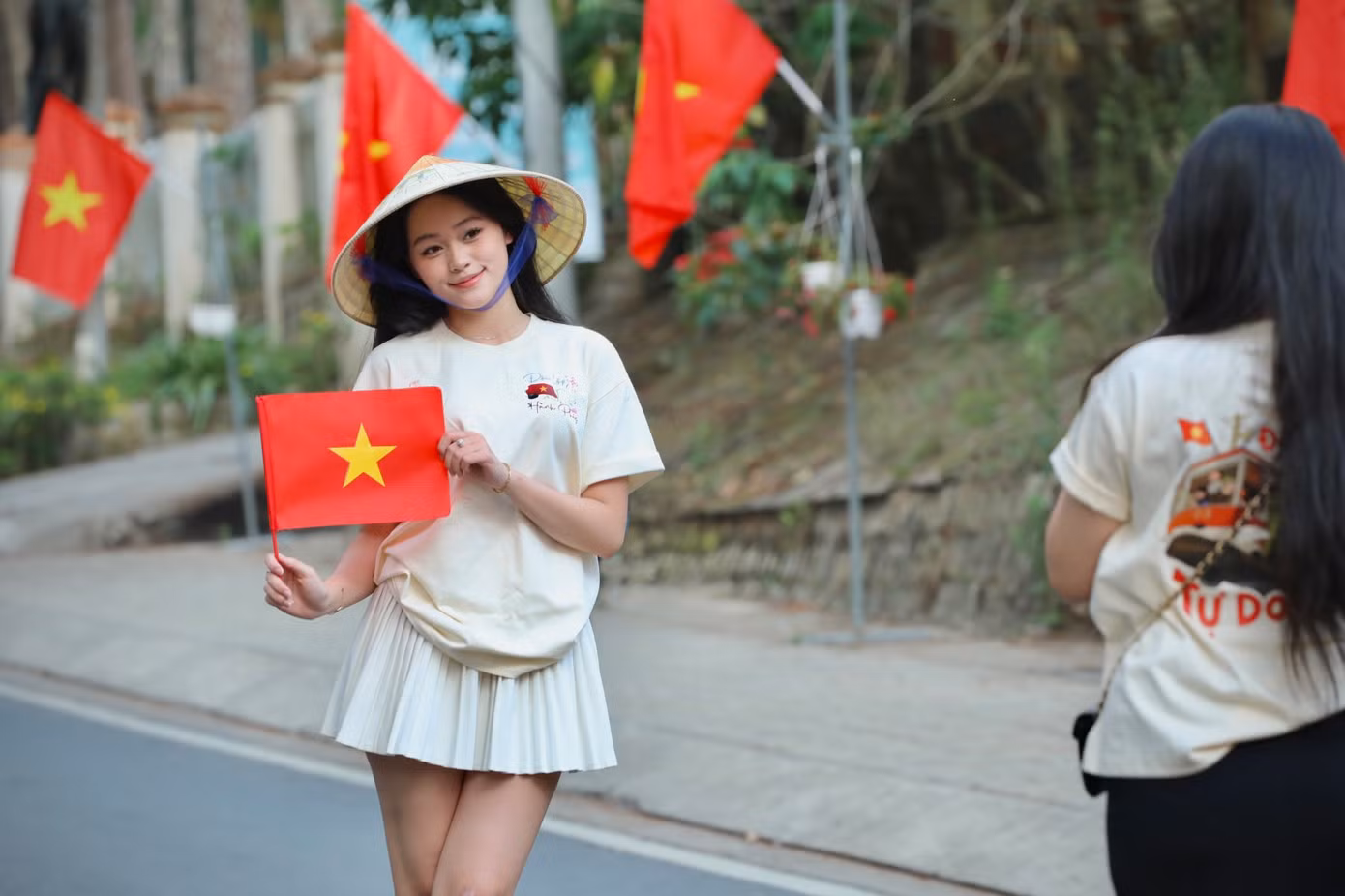 Bạn trẻ khoác lên mình chiếc áo với dòng chữ Độc Lập - Tự Do - Hạnh Phúc và đội nón lá truyền thống check-in con đường cờ hoa.