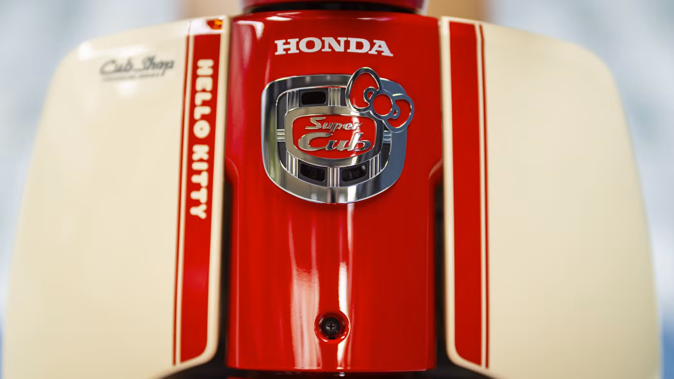 Honda Super Cub Hello Kitty sở hữu phối màu đỏ và be. Logo mèo Hello Kitty xuất hiện trên nhiều vị trí như ốp hông, yếm xe phía trước, bọc yên bằng da, chìa khóa cơ, đồng hồ tốc độ dạng analog kết hợp màn hình kỹ thuật số.