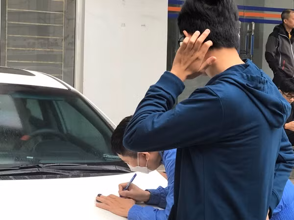 Một phụ huynh (đang viết) lo lắng viết tên tài khoản và mật khẩu của con ra giấy để chạy ra ngoài, đăng nhập rồi lấy giấy báo dự thi và in cho con.