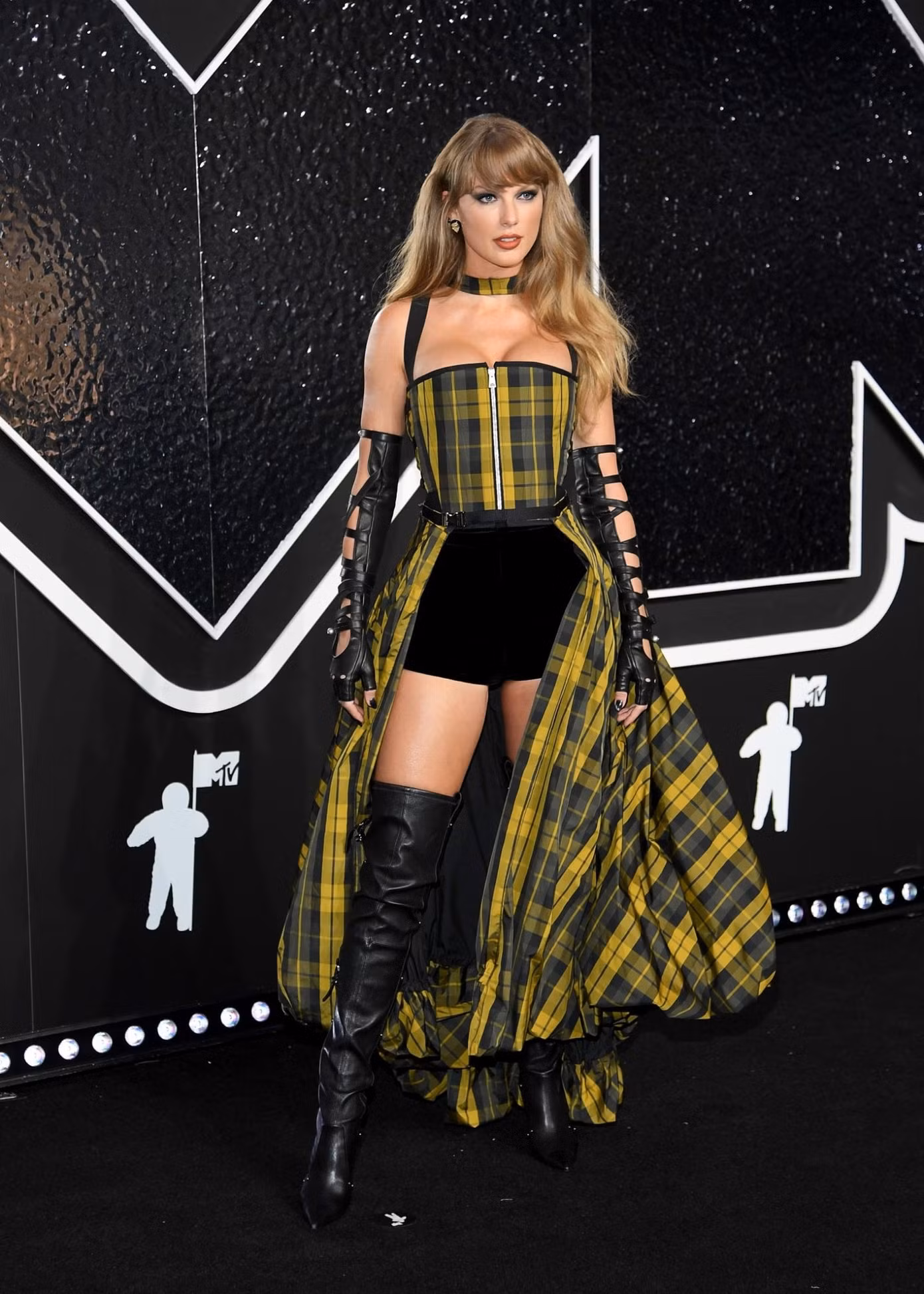 Taylor Swift nổi bật trong chiếc váy corset kẻ sọc vàng đen có dây đeo, kết hợp quần short. Cô tạo vẻ cá tính bằng đôi bốt cao đến đùi và găng tay khoét lỗ.